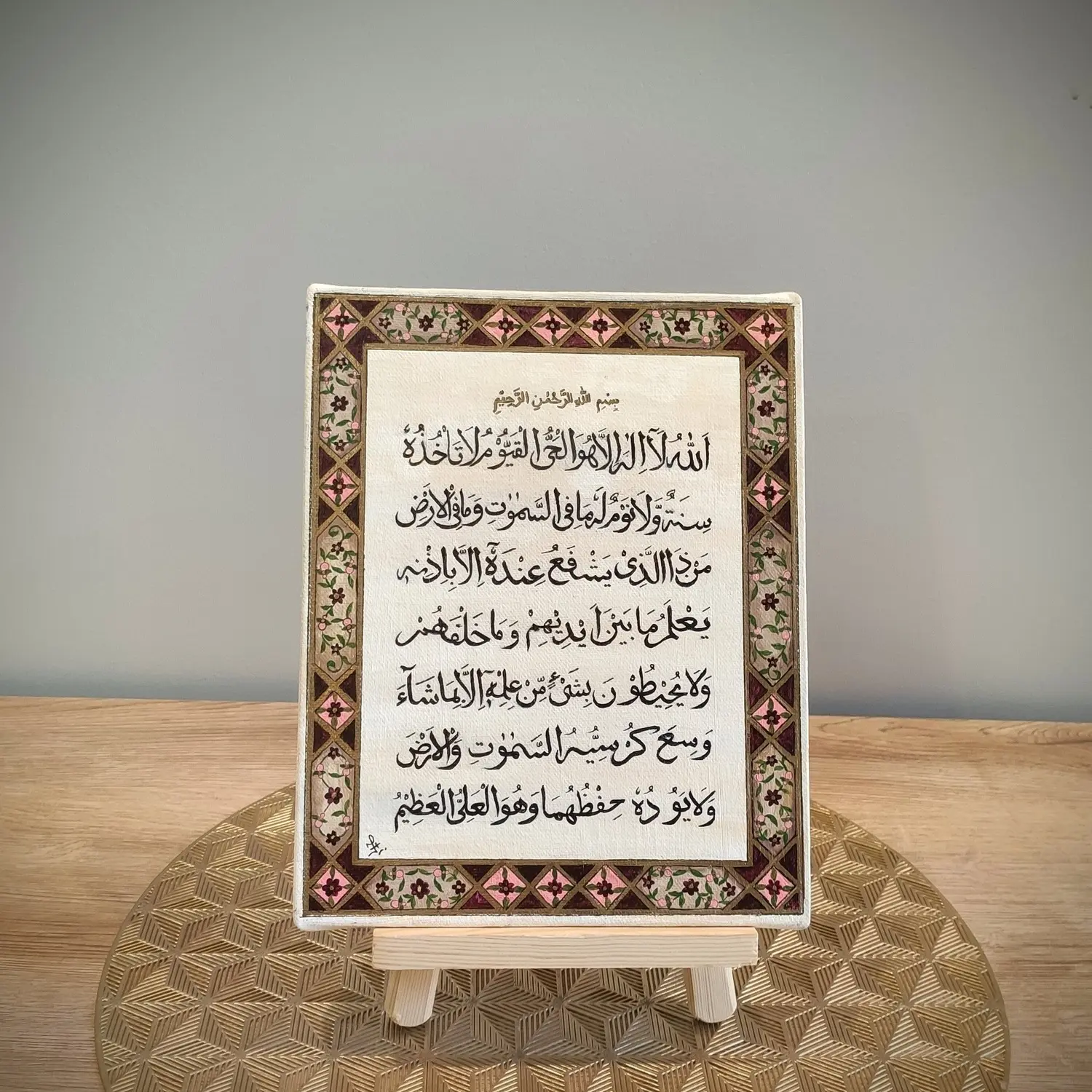 A framed artwork featuring Arabic calligraphy of Ayat al-Kursi is displayed on a small wooden easel. The calligraphy is centered on a cream-colored background, surrounded by an ornate border decorated with floral and geometric patterns in gold, pink, green, and black. The frame and easel rest on a gold geometric-patterned mat atop a wooden surface, against a plain light-colored wall.