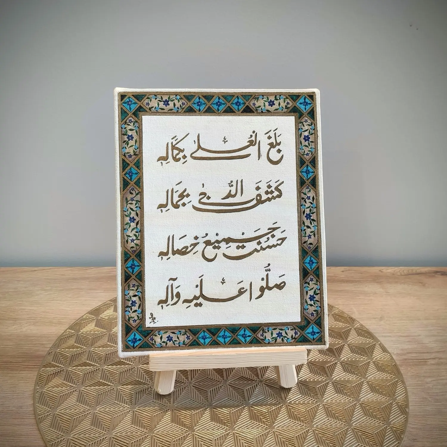 A framed artwork featuring Arabic calligraphy of the verse “Balaghal ‘Ula Bi Kamalihi,” surrounded by a decorative border with turquoise, gold, and floral motifs. The frame is displayed on a small wooden easel placed on a gold geometric-patterned mat against a light background.