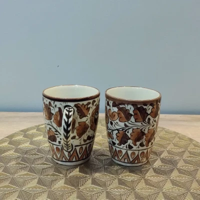Pair of brown Multani pottery mugs with floral patterns, displayed on a gold geometric-patterned mat.