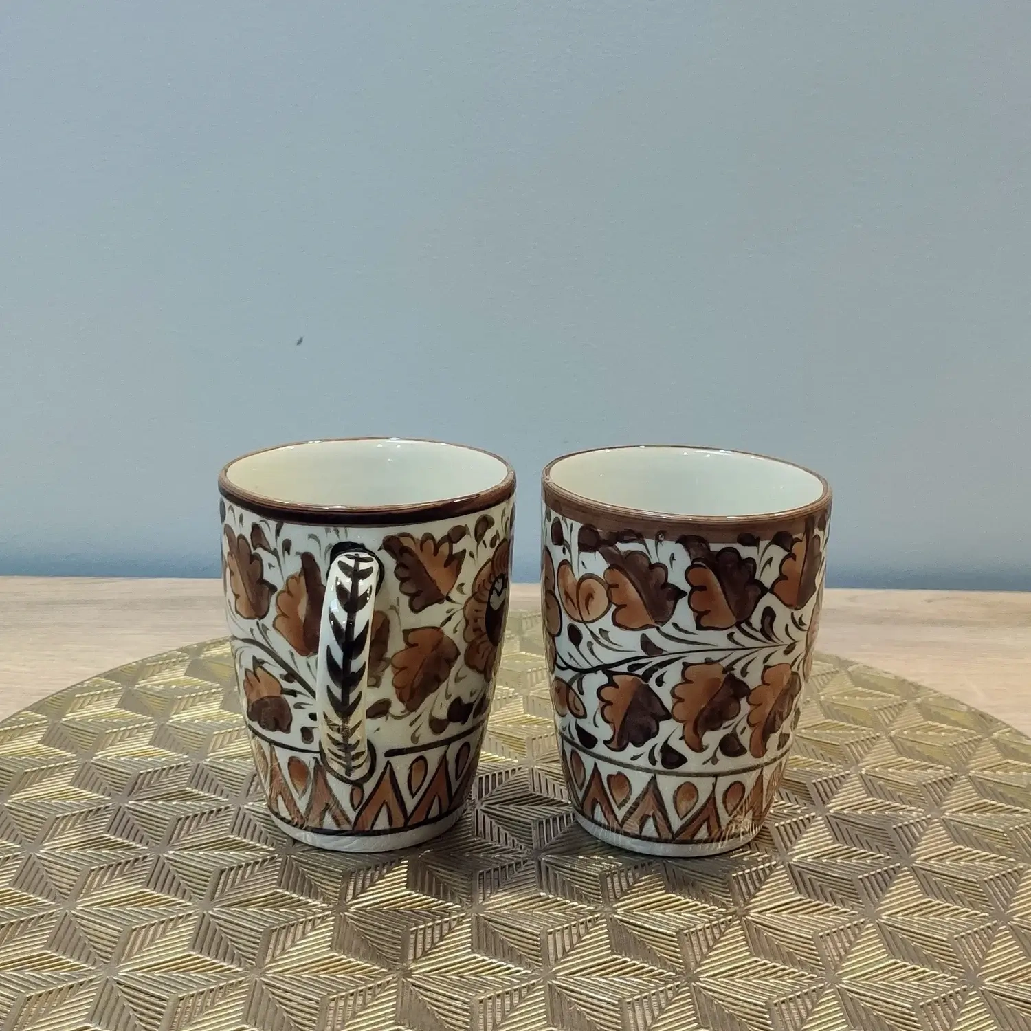 Pair of brown Multani pottery mugs with floral patterns, displayed on a gold geometric-patterned mat.