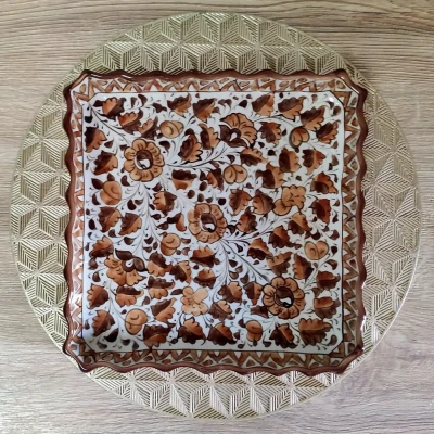 Square brown Multani pottery dish with intricate floral patterns, placed on a gold geometric mat.