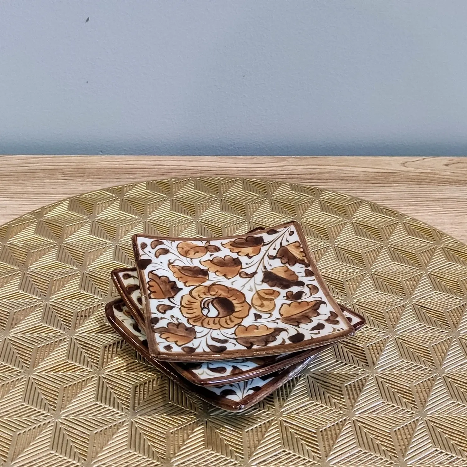 Hand-painted brown Multani pottery square coaster with floral Gul Bale design, placed on a textured golden geometric placemat.