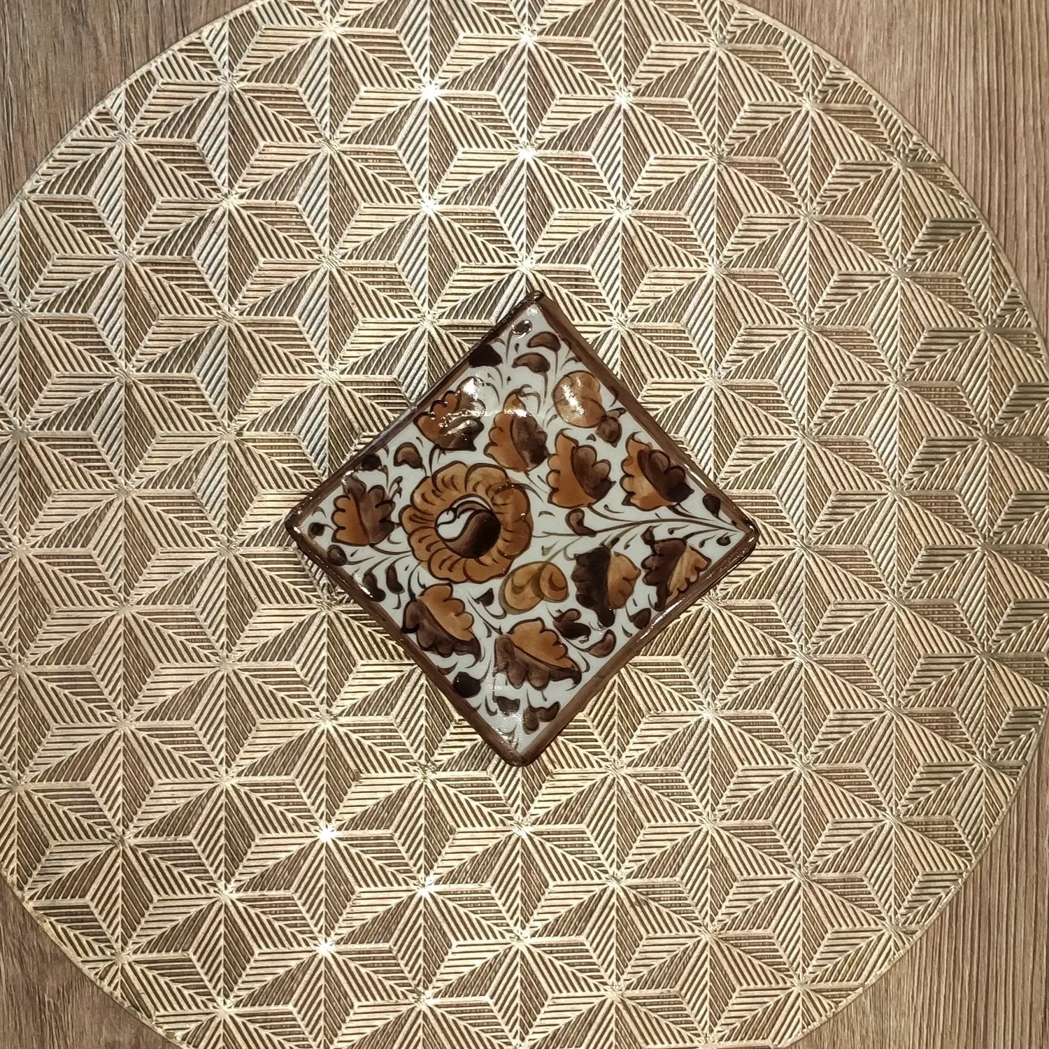 Hand-painted brown Multani pottery square coaster with floral Gul Bale design, placed on a textured golden geometric placemat.