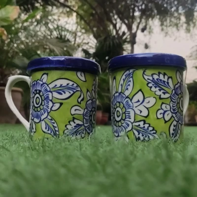 Pair of green Multani pottery mugs with blue and white floral patterns, placed on grass with a garden background.