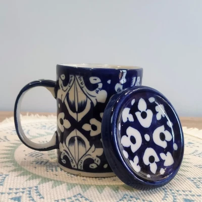 Deep blue Multani pottery mug with white floral motifs and a matching lid, displayed on a patterned woven mat.