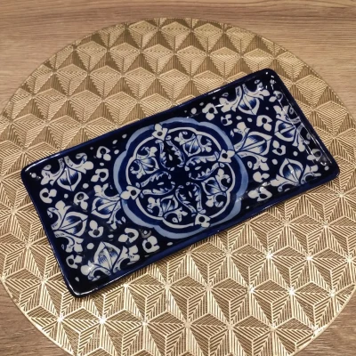 A rectangular Multani blue pottery dish featuring intricate white floral and geometric motifs on a deep blue background, placed on a gold geometric-patterned round mat over a wooden surface.