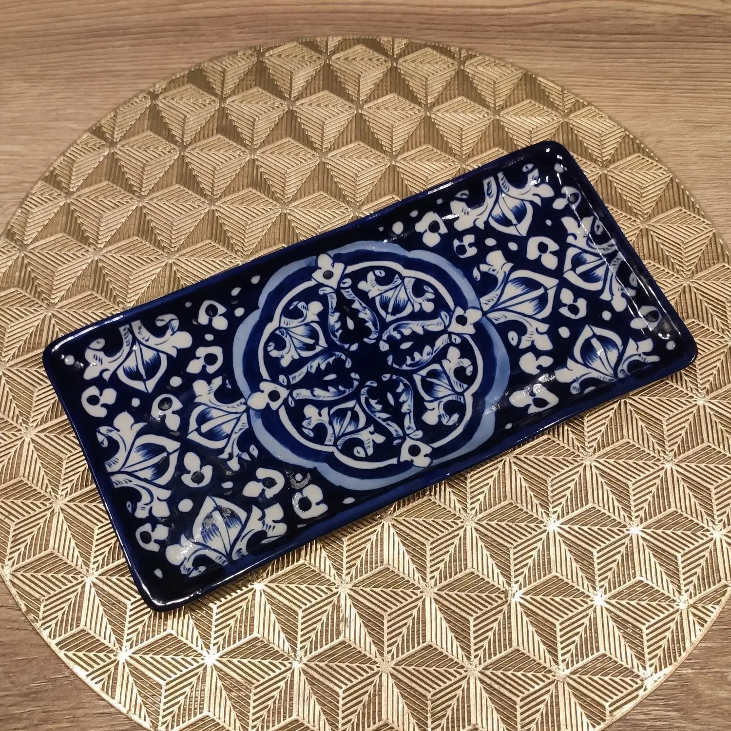 A rectangular Multani blue pottery dish featuring intricate white floral and geometric motifs on a deep blue background, placed on a gold geometric-patterned round mat over a wooden surface.