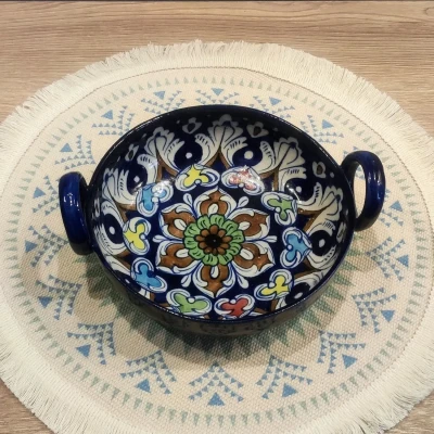 A handcrafted Multani blue pottery bowl with two side handles, featuring intricate floral and geometric patterns in blue, green, brown, and yellow, placed on a round fringed woven mat with light blue tribal motifs.