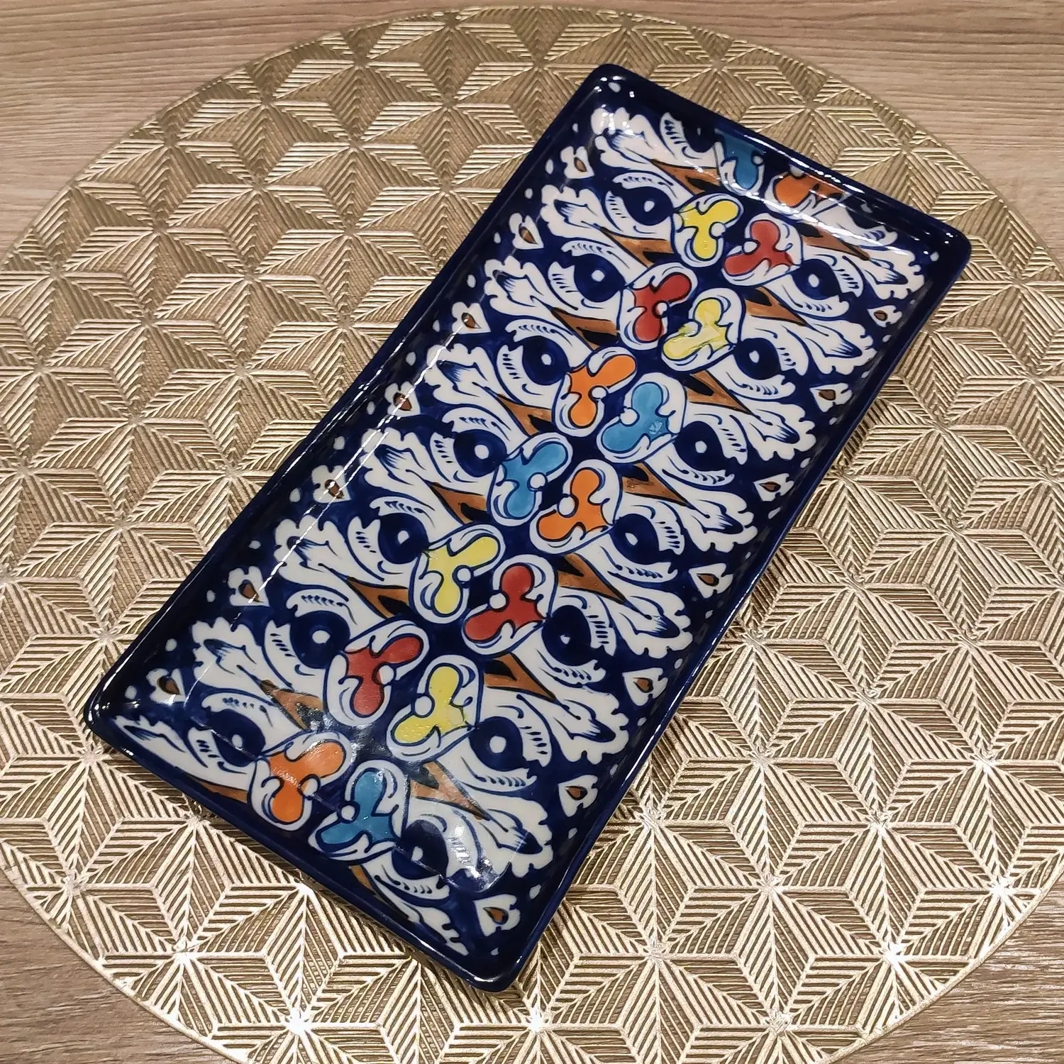 A rectangular Multani pottery dish with vibrant multicolored floral motifs—featuring blue, red, yellow, and orange accents—painted over a deep navy background, placed on a gold geometric-patterned round mat atop a wooden surface.