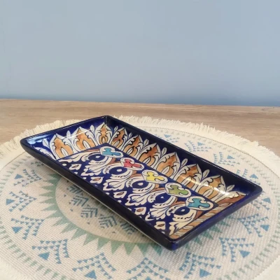 A medium rectangular Multani pottery dish with multicolored floral motifs against a deep blue base, displayed on a round woven mat with teal and blue geometric patterns, set on a wooden surface with a light blue wall in the background.