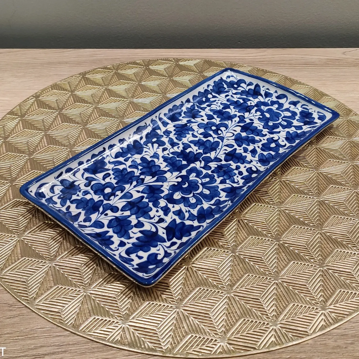 A rectangular Multani blue pottery dish featuring dense hand-painted cobalt floral patterns on a white background, placed on a gold geometric-patterned round mat over a wooden surface.