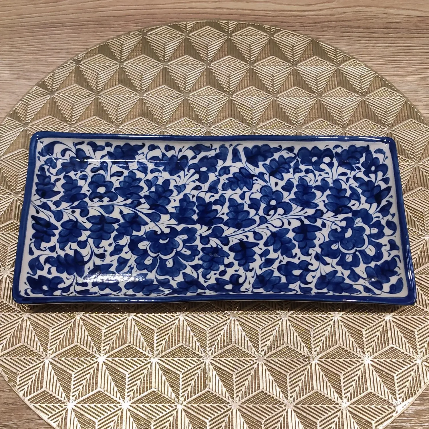A rectangular Multani blue pottery dish featuring dense hand-painted cobalt floral patterns on a white background, placed on a gold geometric-patterned round mat over a wooden surface.