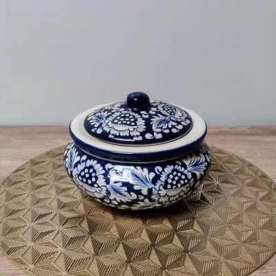 A round ceramic handi-style container with a fitted lid, decorated in dark blue with intricate white floral patterns, sits on a textured gold geometric placemat against a light wall and wooden surface.
