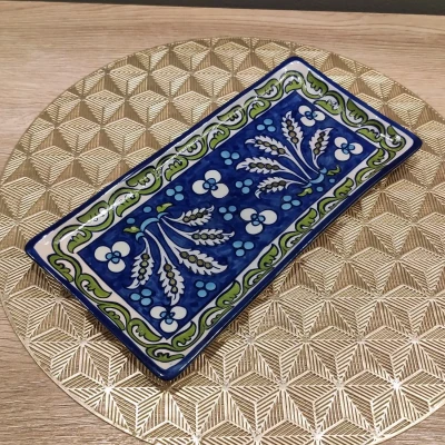 A rectangular Multani pottery dish featuring blue floral and leaf motifs with a green and white decorative border, set against a gold geometric-patterned round mat on a wooden surface.