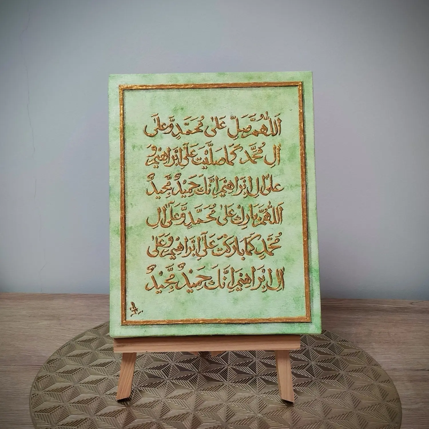 A framed artwork featuring Arabic calligraphy of Durood-e-Ibrahimi written in elegant brown script on a textured green background. The design includes a gold-lined border and is displayed on a wooden easel placed on a decorative gold mat against a light-colored wall.