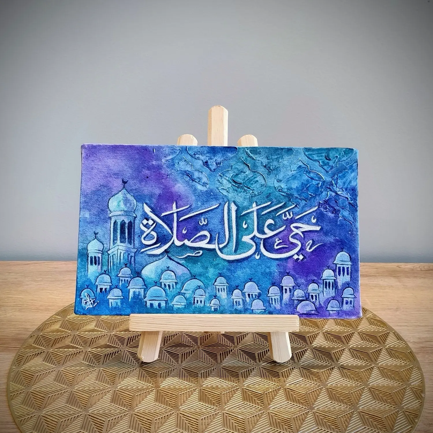 A blue and purple textured canvas featuring the Arabic phrase “Haya Alas Salah” in white calligraphy. A row of painted masjid domes and minarets decorates the bottom. The artwork is displayed on a wooden easel over a gold patterned mat.
