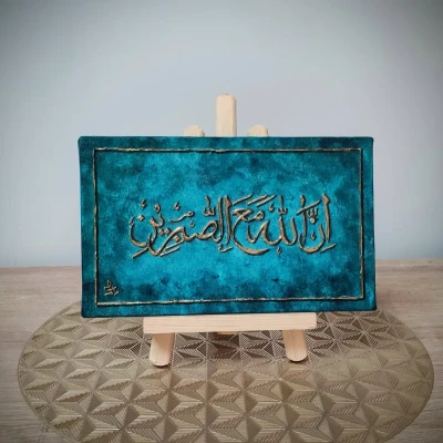 A teal textured calligraphy artwork featuring the Quranic phrase “InnaAllaha Ma’as Sabirin” in raised gold Arabic script. The piece is bordered with a thin gold frame and displayed on a wooden easel over a gold patterned mat.